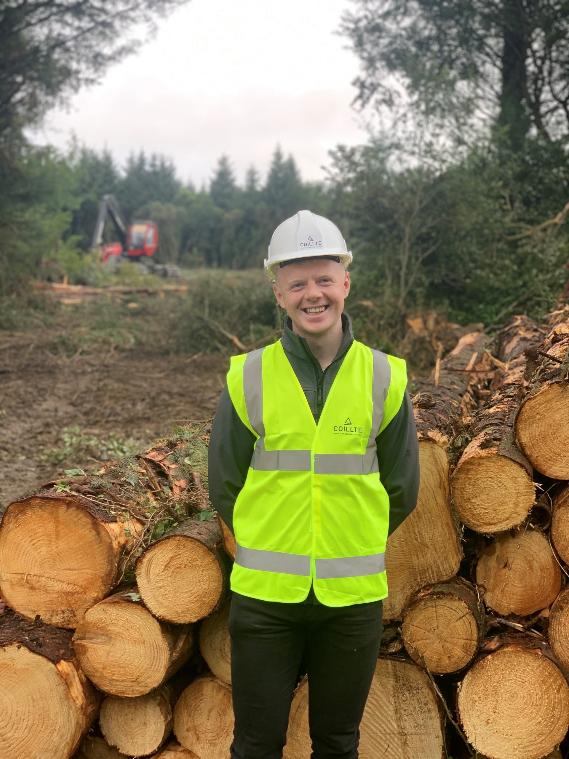 Mark Murphy Coillte Graduate 2022 Picture of Coillte graduate Mark Murphy in high vis jacket and hard hat standing in front of a stack of harvested logs in a forest.