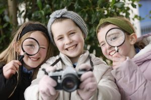 Three young girls holding cameras enthusiastically in front of a tree