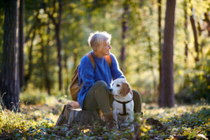 A happy woman with dog on a walk outdoors in forest, resting.