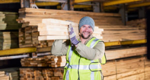 man working at a warehouse at a lumberyard or home improvement store. He is carrying a stack of lumber on his shoulder, smiling at the camera.