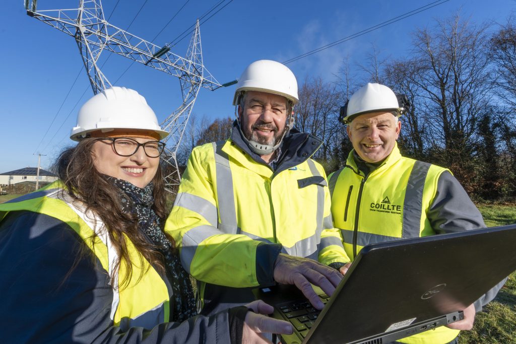 Three people ih hi vis jackets and hard hast holding a laptop computer in front of an electricity pylon close to trees
