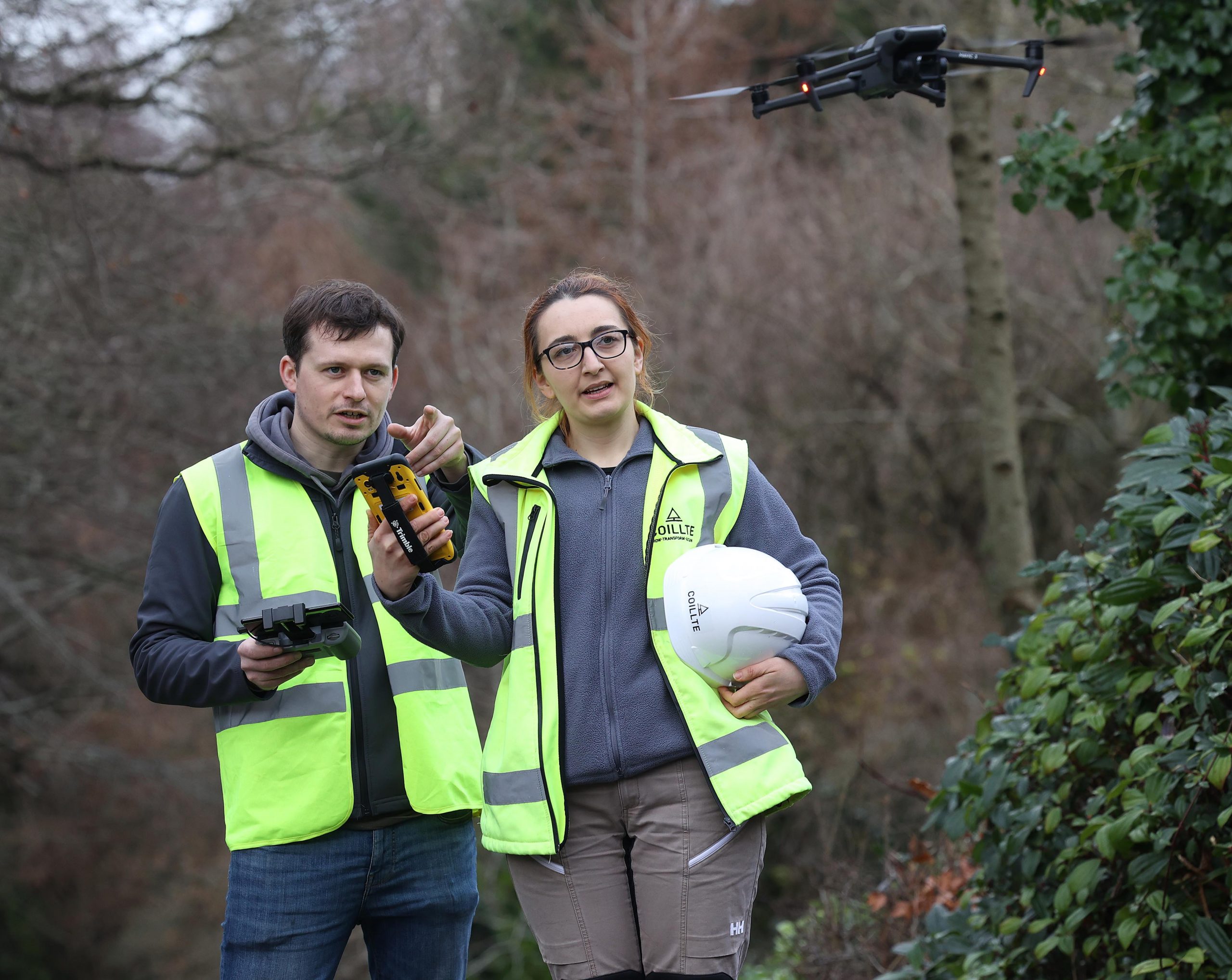 Two foresters standing on a forest road in high visibility jackets operating a drone