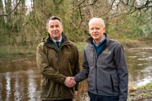 Mark Carlin, Coillte and Barry Fox, Inland Fisherise ireland shaking hands in front of a river flowing through a forested landscape