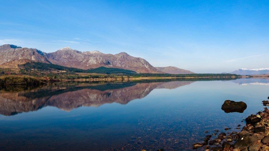 Picture of a forest panted on peatlands at the foot of a mountain taken from across a lake on a sunny day with clear blue skies