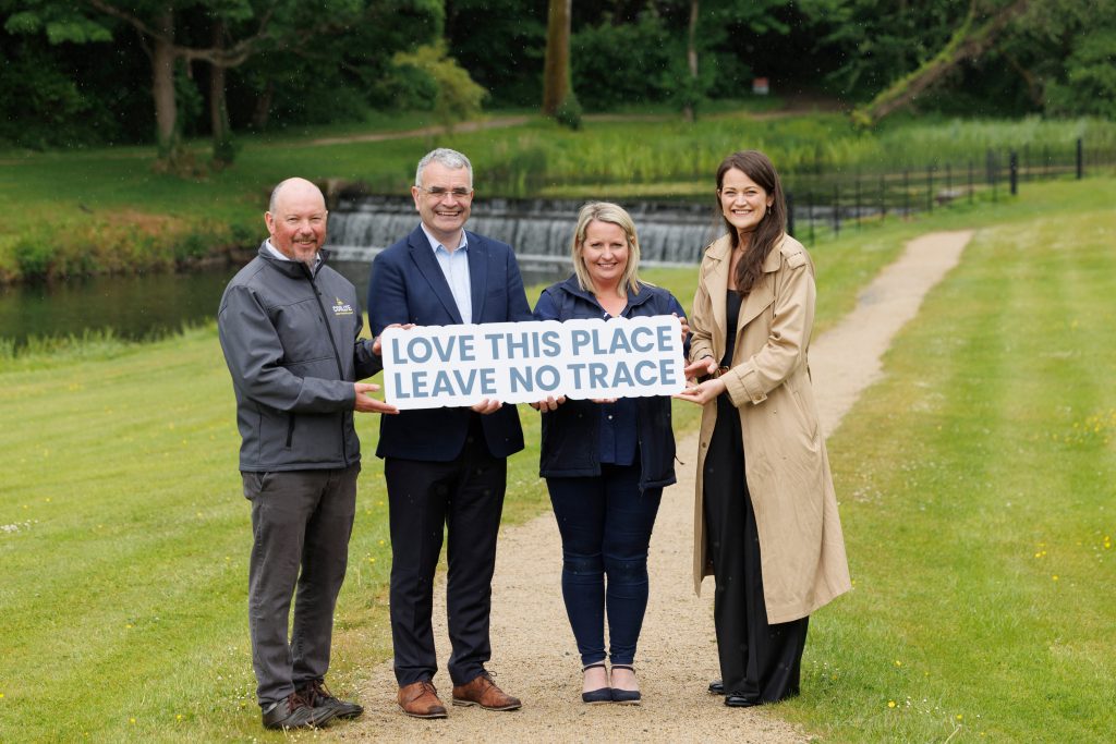Four people holding a love this place leave no trace sign on a pathway surrounded by green lawn